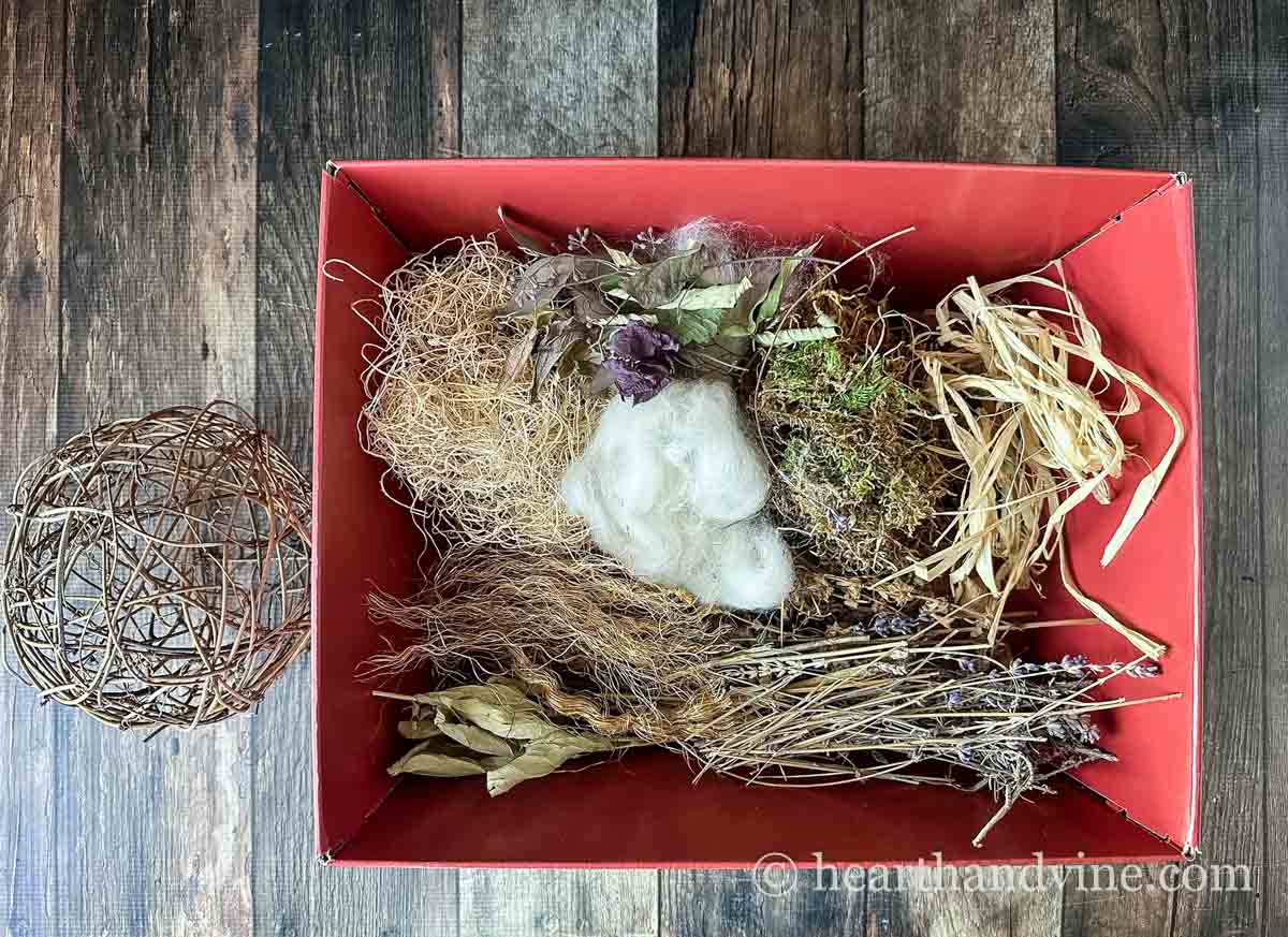 Supplies to create a nesting materials for birds ball including a grapevine ball, and a box with supplies including, dried lavender, wool, moss, excelsior, and sisal.