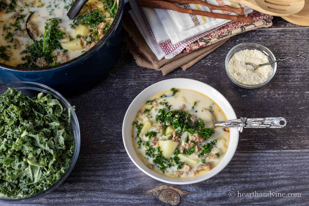A bowl of zuppa toscana next to a smaller bowl of Parmesan cheese, a bowl of kale and a pot of the same soup.