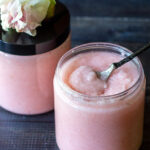 Two jars of blush sugar scrub with the front one open and a small spoon in it.