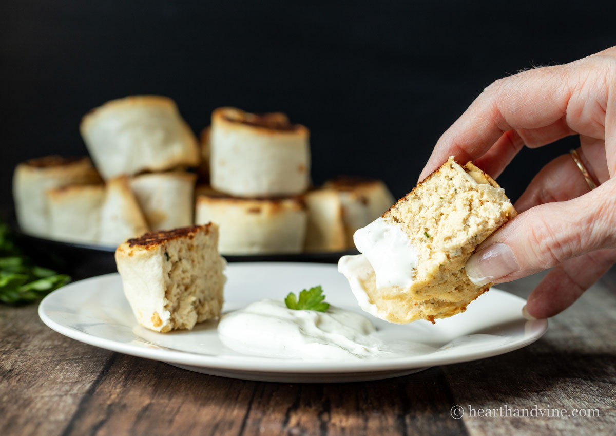 A hand holding half of a chicken pinwheel dipping into the ranch sauce above a plate with the sauce and the other half.