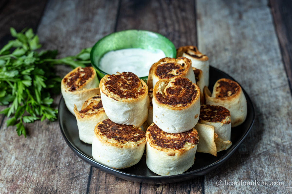 Chicken pinwheels stacked on a black plate with a small bowl of dipping sauce.