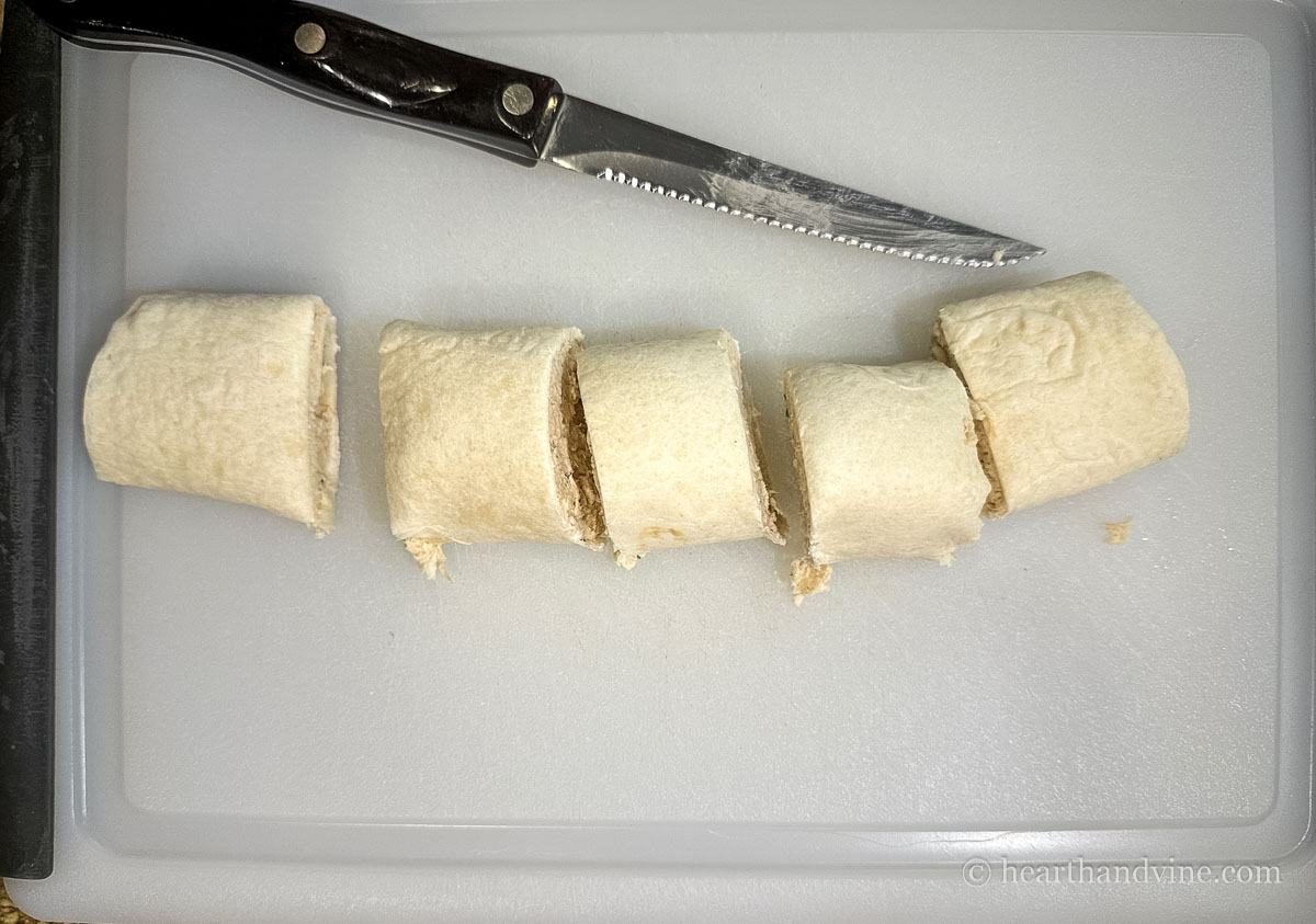 A knife on a cutting board with cut pieces of chicken tortilla roll.
