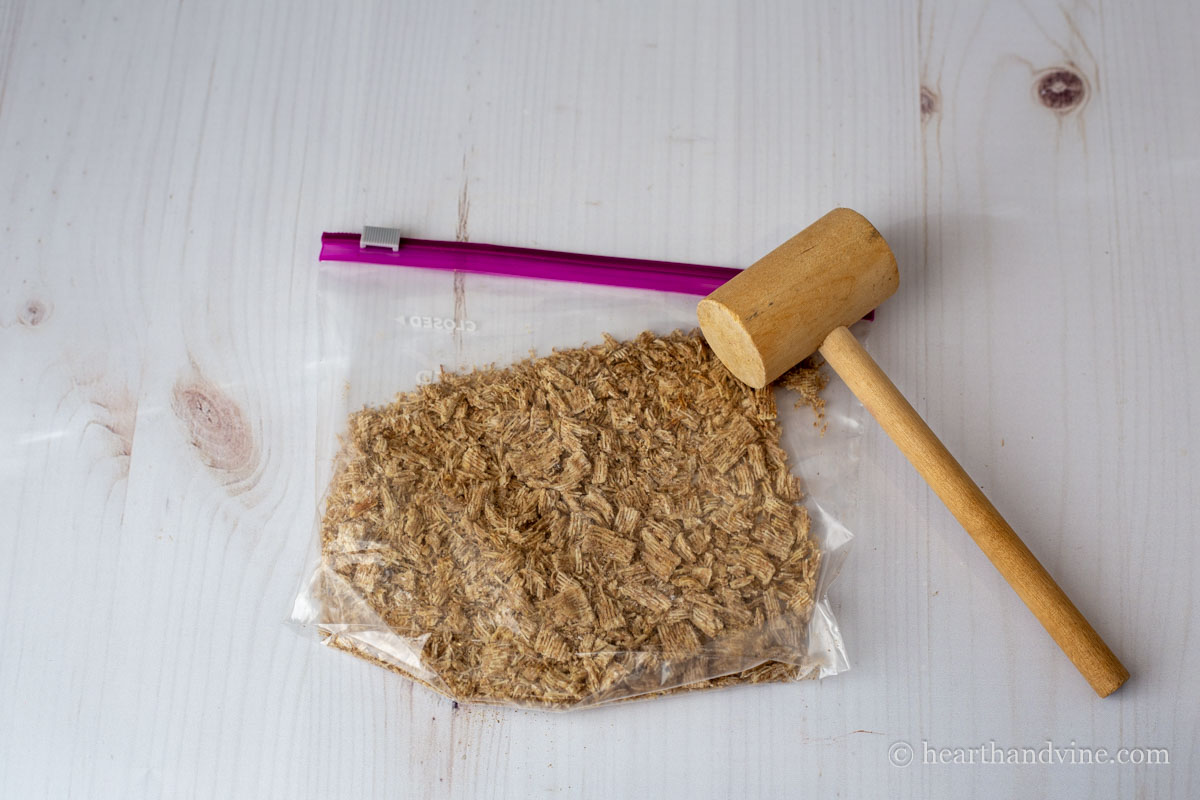 Shredded wheat cereal crushed in a plastic bag with a wooden mallet.