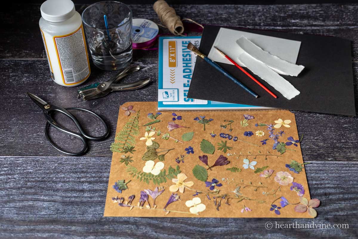 Supplies for pressed flower bookmarks including pressed flowers, scissors, laminating sheets, mod podge, heavy paper, paintbrushes and ribbon.