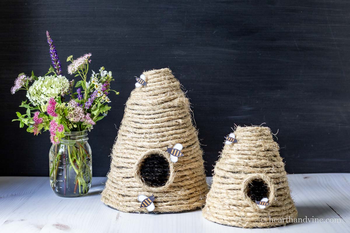 A large and small bee skep with paper bees next to a mason jar filled with flowers.
