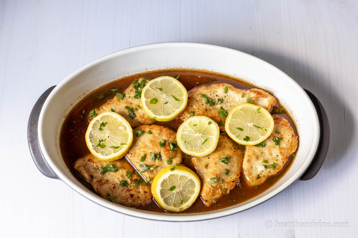 Chicken piccata in a serving dis with fresh parsley and lemon slices on top.