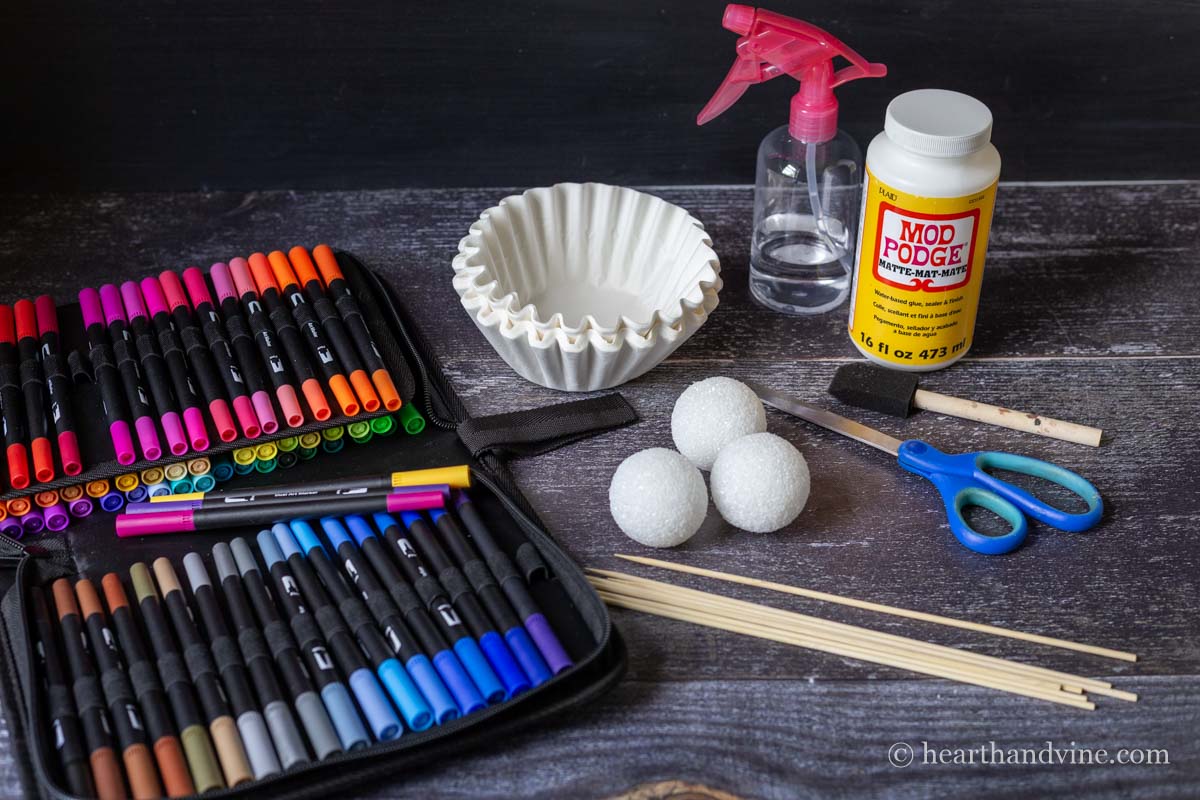 Supplies for coffee filter flowers including markers, foam balls, scissors, mod podge, skewers and a spray bottle with water.