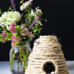A small DIY bee skep with paper bees and a glass of flowers.