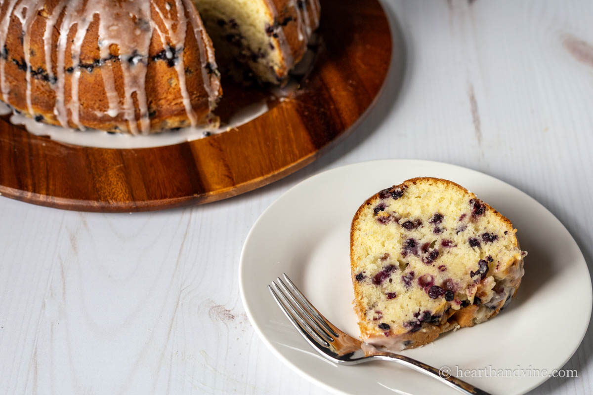 Slice of blueberry and lemon bundt cake on a white plate with a fork next to the entire cake.