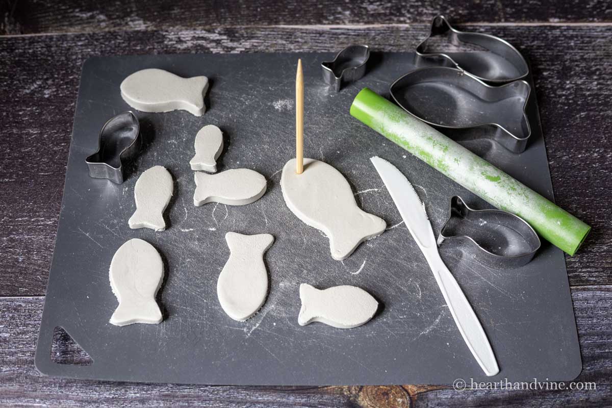 A wooden skewer pressed into an air dry clay fish in the eye are surrounded by more fish, fish cookie cutters and a plastic knife on a plastic mat.