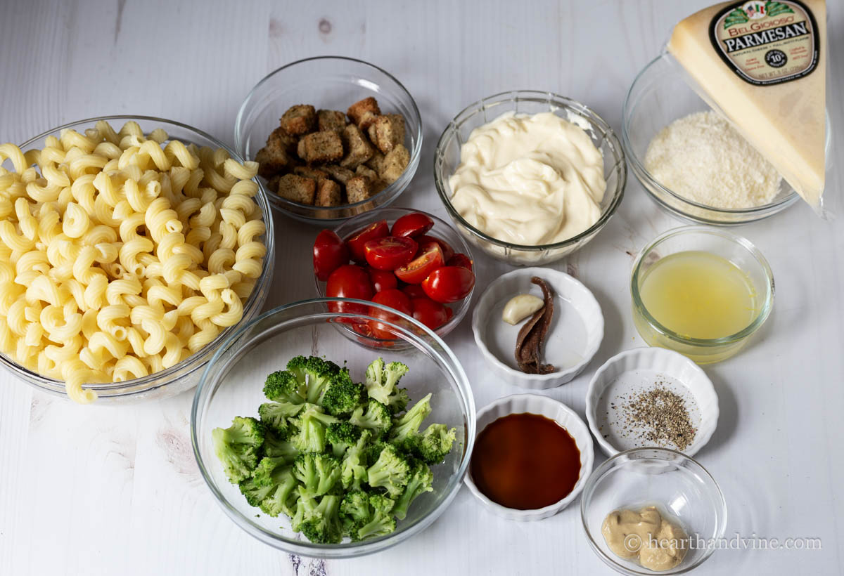 Ingredients for a Caesar pasta salad including pasta, broccoli, tomatoes, mayo, lemon juice, cheese, anchovies, garlic, mustard and worcestershire.