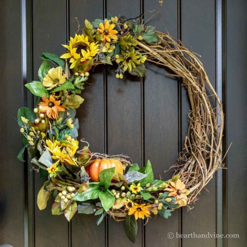 Grapevine wreath partially decorated with sunflowers, a small pumpkin and other green foliage.
