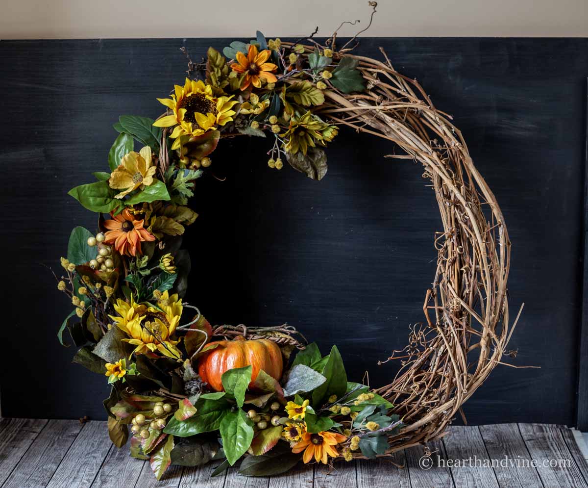 A small pumpkin nested in a grapevine wreath with sunflowers and greenery set on a top leaning on a wall.