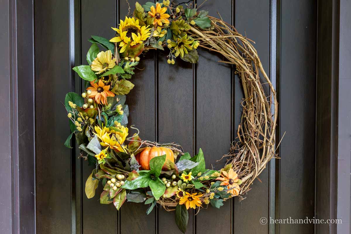 A small pumpkin nested in a grapevine wreath with sunflowers and greenery on a brown front door.