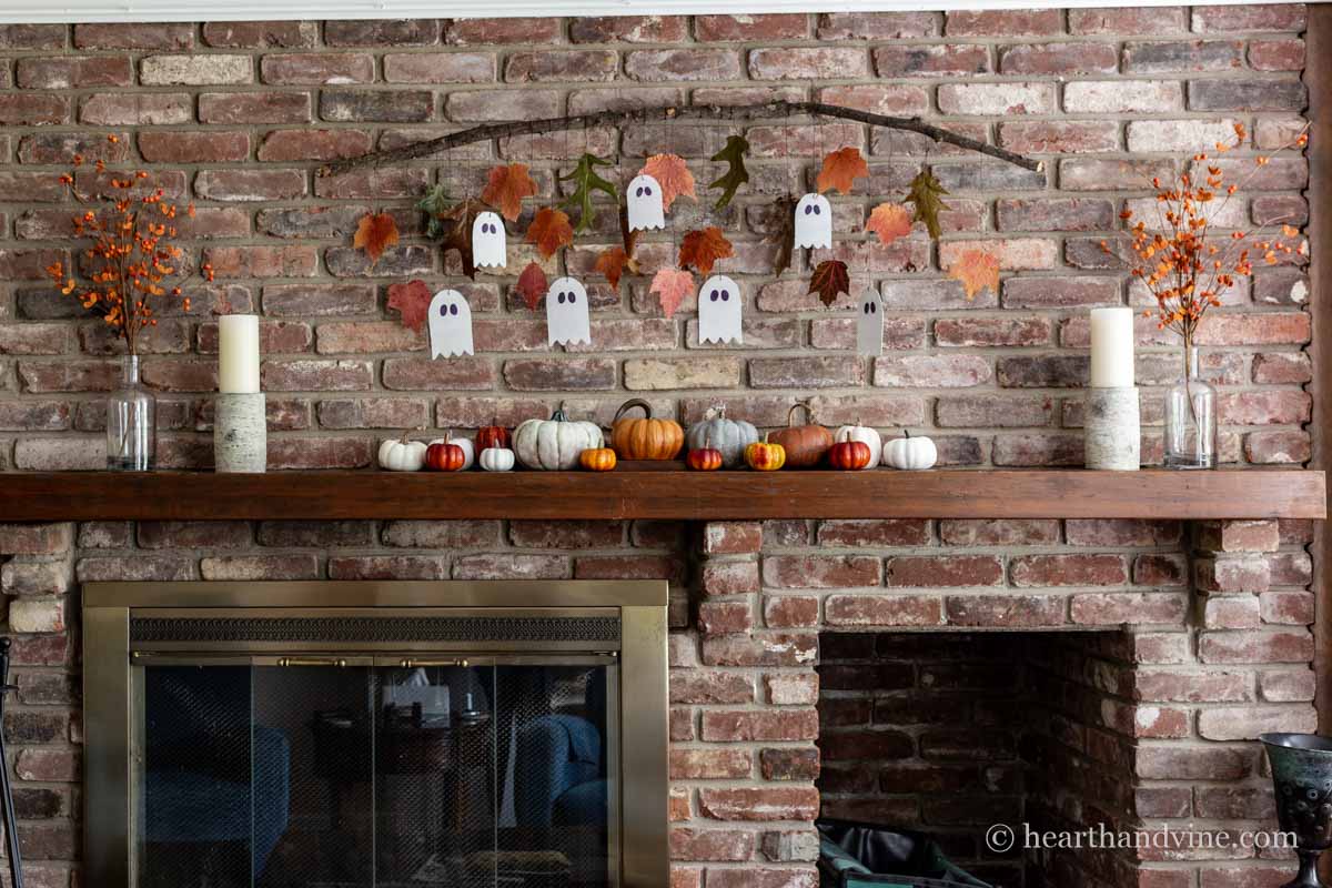 View of full fall decorated fireplace and mantle with ghost garland and leaves, faux pumpkins, white candles and faux orange berries in clear bottls.