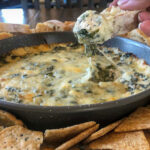 A hand lifting a taste of a cream cheese spinach dip on a bread round.