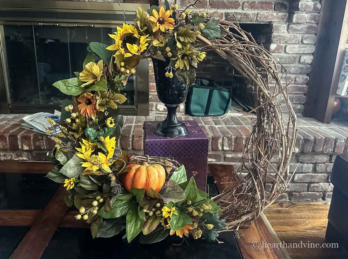 A sunflower and pumpkin wreath propped up on a box and urn on a table.