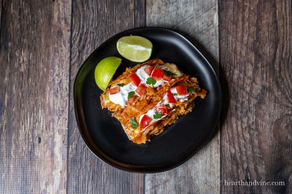 Two baked chicken tacos on a plate with sour cream, tomatoes and cilantro next to lime wedges.