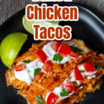 Two baked chicken tacos on a plate with lime wedges and topped with sour cream, tomatoes and cilantro.